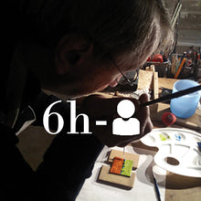 Charger l&#39;image dans la galerie, Homme pendant l&#39;atelier de découverte, concentration sur sa pièce unique en émail, assis au plan de travail, tient un pinceau dans sa main.
