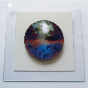 Cette décoration murale ronde, en emaux joaillier grand feu avec de forte présence de la couleur bleu et du rouge, rouille sombre est sur un encadrement blanc.