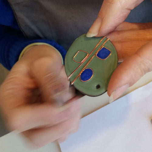 Pièce unique ovale en émail vert avec deux petits carrés bleus tenu par les mains d'une femme.
